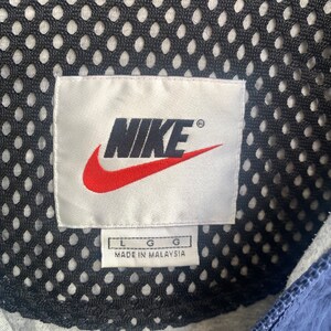 May include: A white tag with the Nike logo in black and red, with the text "NIKE" and a registered trademark symbol. The tag also has the text "L G G" and "MADE IN MALAYSIA".