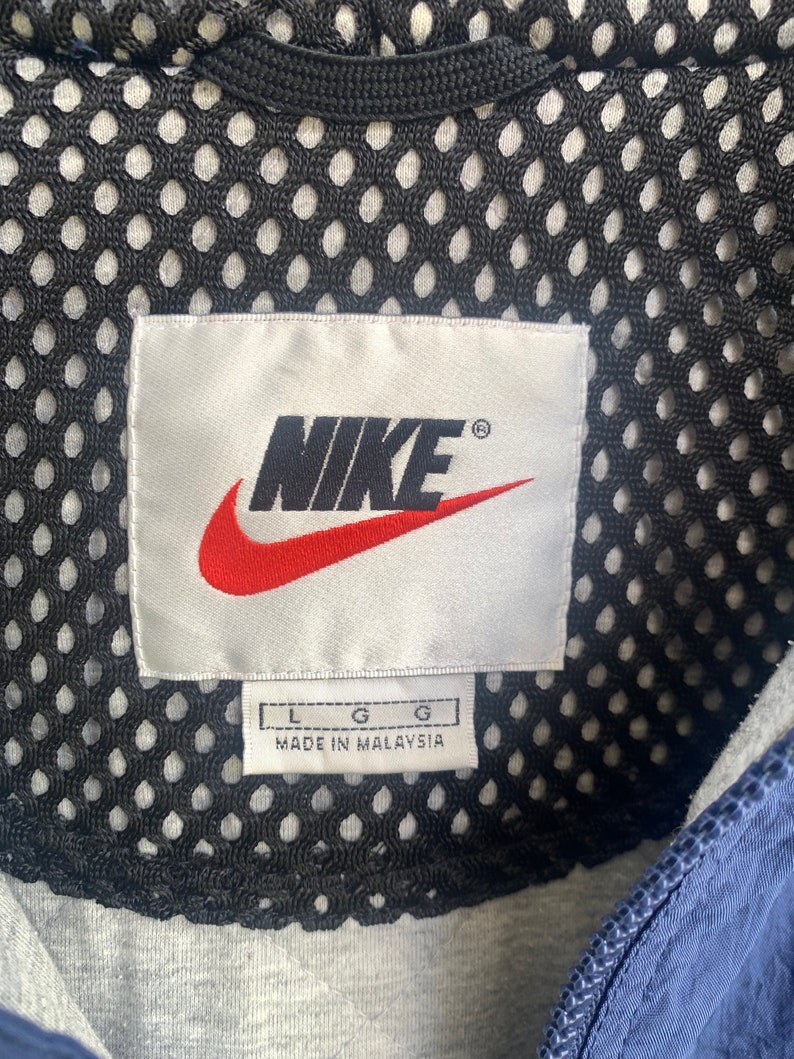 May include: A white tag with the Nike logo in black and red, with the text "NIKE" and a registered trademark symbol. The tag also has the text "L G G" and "MADE IN MALAYSIA".