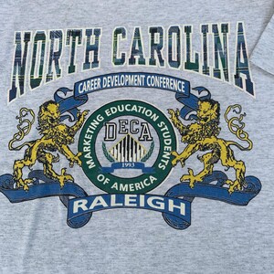 Pu&ograve; includere: T-shirt grigio melange con la scritta "NORTH CAROLINA" in un motivo a quadri scozzesi. Sotto, un emblema circolare con la scritta "DECA 1993" con "Career Development Conference" e "RALEIGH". Due leoni dorati fiancheggiano l'emblema.