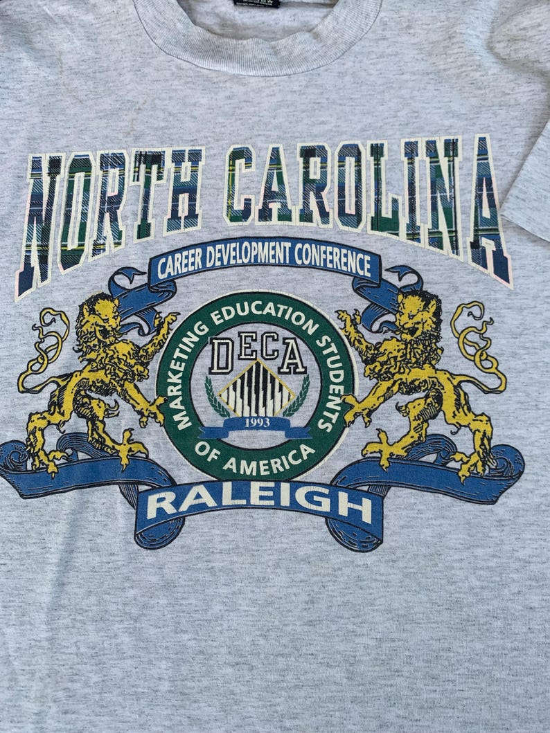Pu&ograve; includere: T-shirt grigio melange con la scritta "NORTH CAROLINA" in un motivo a quadri scozzesi. Sotto, un emblema circolare con la scritta "DECA 1993" con "Career Development Conference" e "RALEIGH". Due leoni dorati fiancheggiano l'emblema.