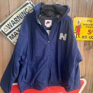 May include: A navy blue hooded jacket with a white Nike logo and a white "N" on the front. The jacket has a mesh lining and a zip closure.