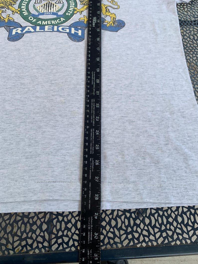Pu&ograve; includere: Una maglietta grigio chiaro con una grafica con la scritta "DECA" e "MARKETING STUDENTS OF AMERICA RALEIGH 1993". Un righello nero &egrave; posto verticalmente sulla maglietta, misurando circa 76 cm.