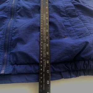 May include: A navy blue windbreaker jacket with a zipper closure and a pocket on the right side. The jacket has a ribbed hem and cuffs.