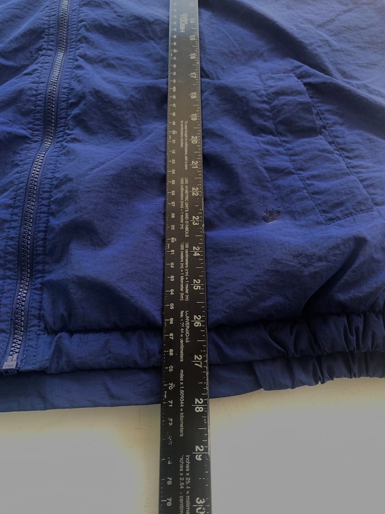 May include: A navy blue windbreaker jacket with a zipper closure and a pocket on the right side. The jacket has a ribbed hem and cuffs.