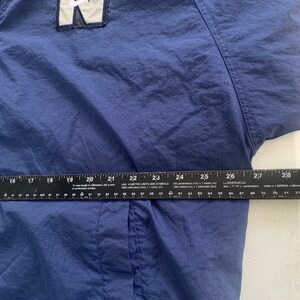 May include: A navy blue windbreaker jacket with a white and black Nike logo patch on the front. The jacket has a zip fastening and a drawstring hood.