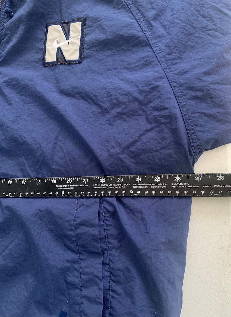 May include: A navy blue windbreaker jacket with a white and black Nike logo patch on the front. The jacket has a zip fastening and a drawstring hood.