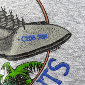 Puede incluir: Sudadera gris con un gr&aacute;fico que presenta un barco, palmeras y el texto "CLUB SUN" en azul. El dise&ntilde;o est&aacute; enmarcado por un c&iacute;rculo naranja y azul, con la palabra "NIGHTS" en azul. La firma del artista "Anderson" es visible.