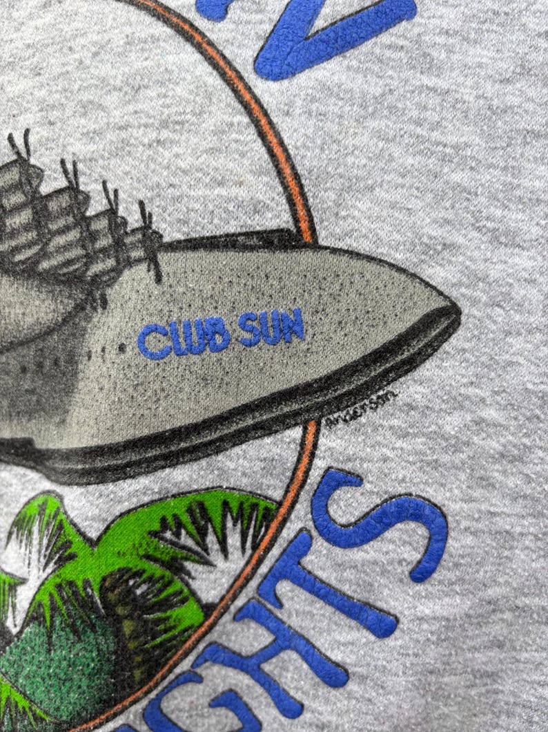 Puede incluir: Sudadera gris con un gr&aacute;fico que presenta un barco, palmeras y el texto "CLUB SUN" en azul. El dise&ntilde;o est&aacute; enmarcado por un c&iacute;rculo naranja y azul, con la palabra "NIGHTS" en azul. La firma del artista "Anderson" es visible.