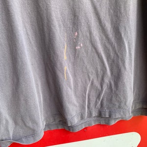 May include: A close-up of a grey t-shirt with a light brown stain and a pink stain. The t-shirt is folded and the bottom edge is visible.