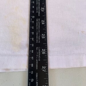 May include: A black ruler with white markings and the words "LINS AR METRIC UNITS AND SYMBOLS" and "CONVERSIONS" printed on it. The ruler measures up to 28 inches or 71 centimeters.