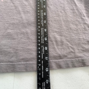 May include: A grey t-shirt with a seam along the bottom edge. A ruler is placed next to the shirt, showing the length of the shirt is 73.66 cm.