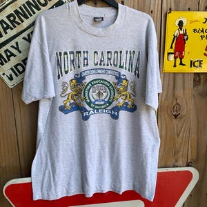 Pu&ograve; includere: Una maglietta grigio melange con la scritta "NORTH CAROLINA" in verde e blu. Sotto, le parole "CAREER DEVELOPMENT CONFERENCE" e "DECA" con due grafiche di leoni. La maglietta &egrave; appesa a una gruccia nera.