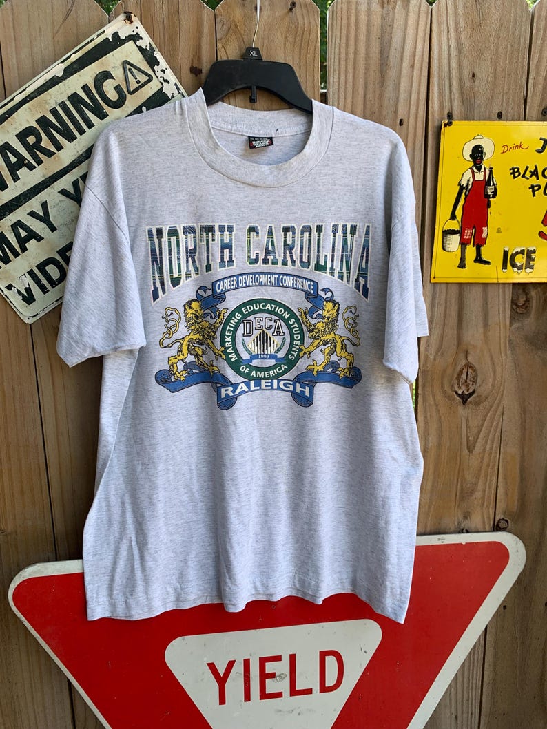 Pu&ograve; includere: Una maglietta grigio melange con la scritta "NORTH CAROLINA" in verde e blu. Sotto, le parole "CAREER DEVELOPMENT CONFERENCE" e "DECA" con due grafiche di leoni. La maglietta &egrave; appesa a una gruccia nera.