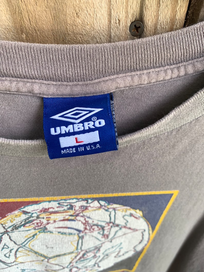 May include: A grey t-shirt with a blue and white Umbro logo and the text "MADE IN U.S.A." The shirt features a graphic of a globe with a yellow outline.