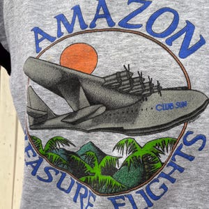 Puede incluir: Sudadera gris con un gr&aacute;fico vintage de un hidroavi&oacute;n y el texto "AMAZON TREASURE FLIGHTS" en azul. El dise&ntilde;o incluye un sol naranja, palmeras verdes y monta&ntilde;as, evocando un tema de viaje retro.