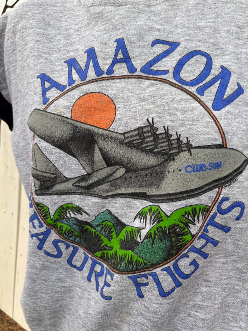Puede incluir: Sudadera gris con un gr&aacute;fico vintage de un hidroavi&oacute;n y el texto "AMAZON TREASURE FLIGHTS" en azul. El dise&ntilde;o incluye un sol naranja, palmeras verdes y monta&ntilde;as, evocando un tema de viaje retro.