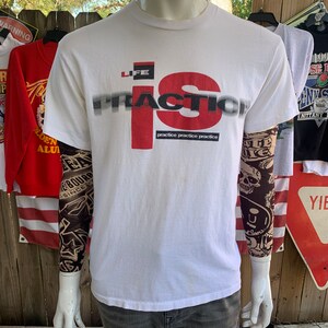 May include: White t-shirt with a red and black graphic that says "Life is Practice" with the word "Practice" repeated three times below the graphic.