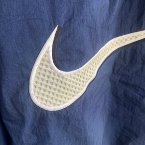 May include: A navy blue jacket with a white and black Nike swoosh logo. The swoosh is made of a mesh material.