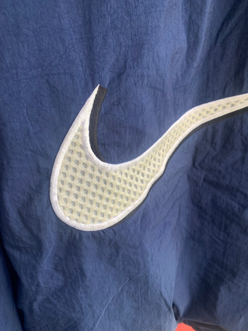 May include: A navy blue jacket with a white and black Nike swoosh logo. The swoosh is made of a mesh material.