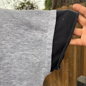 Puede incluir: Primer plano de una manga de camiseta gris con una manga negra debajo. La tela gris tiene un aspecto texturizado. El borde de la manga negra es visible, creando un efecto en capas. El fondo est&aacute; borroso.