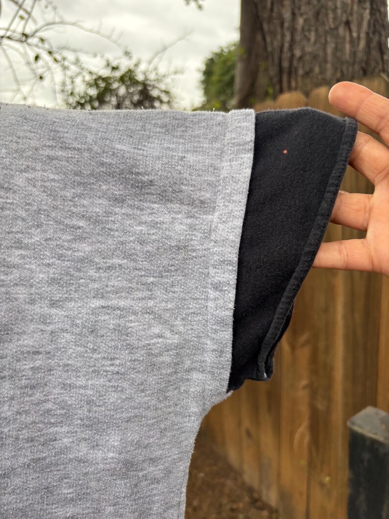 Puede incluir: Primer plano de una manga de camiseta gris con una manga negra debajo. La tela gris tiene un aspecto texturizado. El borde de la manga negra es visible, creando un efecto en capas. El fondo est&aacute; borroso.