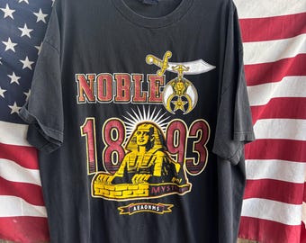 May include: Black t-shirt with a graphic featuring the word "NOBLE" in red and gold, the numbers "1893", and an illustration of the Sphinx. The shirt also displays a sword and crescent symbol, and the text "MYSTIC" and "AEAONMS".