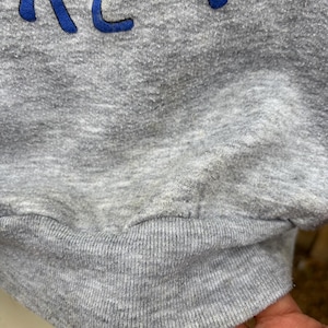 Puede incluir: Primer plano de una sudadera gris con el texto azul "ARE YOU". La sudadera tiene un dobladillo y pu&ntilde;os acanalados. La tela parece ser un material suave y afelpado.