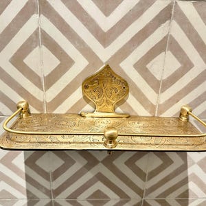 Puede incluir: Un estante de metal dorado con intrincados dise&ntilde;os florales y de volutas. El estante tiene una base rectangular, una placa trasera decorativa y dos brazos curvos. Est&aacute; montado en una pared de azulejos estampados.