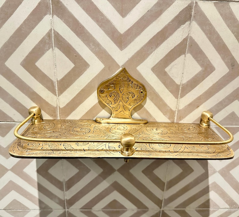 Puede incluir: Un estante de metal dorado con intrincados dise&ntilde;os florales y de volutas. El estante tiene una base rectangular, una placa trasera decorativa y dos brazos curvos. Est&aacute; montado en una pared de azulejos estampados.