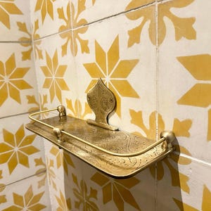 Puede incluir: Un estante de metal dorado con un dise&ntilde;o ornamentado, montado en una pared con azulejos estampados amarillos y blancos. El estante tiene un frente curvo y un soporte trasero decorativo.
