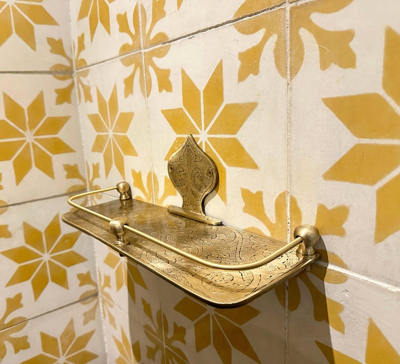 Puede incluir: Un estante de metal dorado con un dise&ntilde;o ornamentado, montado en una pared con azulejos estampados amarillos y blancos. El estante tiene un frente curvo y un soporte trasero decorativo.