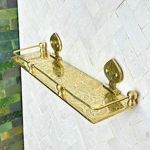 Puede incluir: Una estanter&iacute;a de pared rectangular dorada con un intrincado patr&oacute;n floral grabado. La estanter&iacute;a tiene un borde frontal elevado y est&aacute; sostenida por soportes decorativos en forma de hoja. La estanter&iacute;a est&aacute; montada en una pared de azulejos blancos con dise&ntilde;o de espiga.