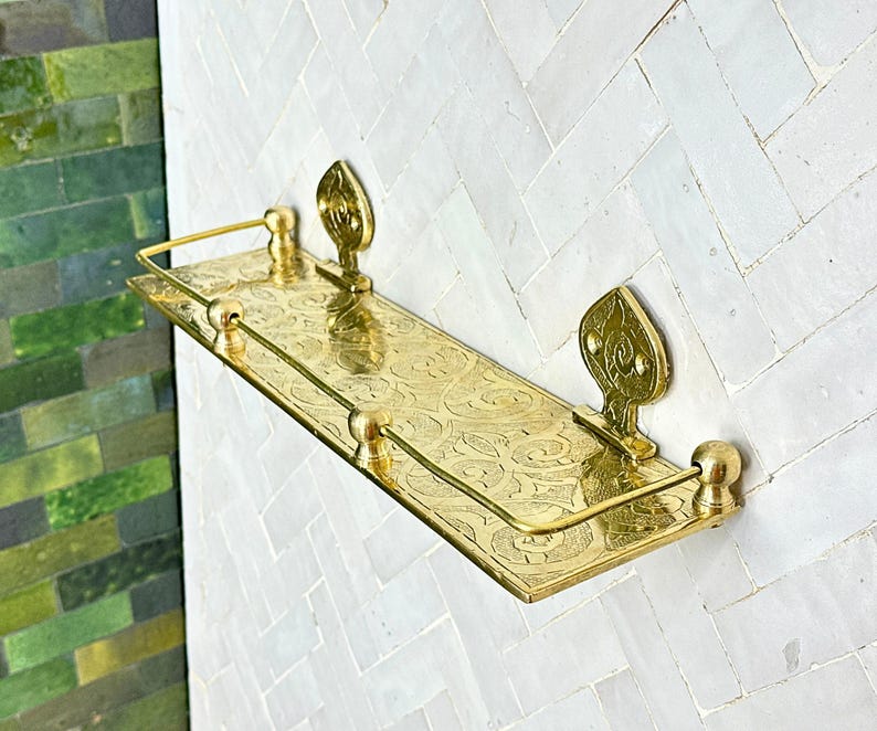 Puede incluir: Una estanter&iacute;a de pared rectangular dorada con un intrincado patr&oacute;n floral grabado. La estanter&iacute;a tiene un borde frontal elevado y est&aacute; sostenida por soportes decorativos en forma de hoja. La estanter&iacute;a est&aacute; montada en una pared de azulejos blancos con dise&ntilde;o de espiga.
