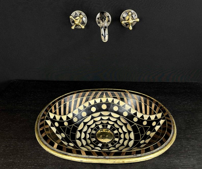 K&ouml;nnte beinhalten: Ein ovales Waschbecken in Schwarz und Wei&szlig; mit einem geometrischen Muster. Das Waschbecken hat einen Messingrand und ist vor einem schwarzen Hintergrund platziert. An der Wand &uuml;ber dem Waschbecken befinden sich zwei Messingkn&ouml;pfe.