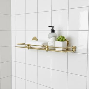 Puede incluir: Estante de ba&ntilde;o de metal dorado montado en azulejos blancos. El estante contiene una toalla blanca enrollada, una botella de jab&oacute;n y una peque&ntilde;a planta suculenta en maceta. El estante tiene detalles decorativos y una barra.
