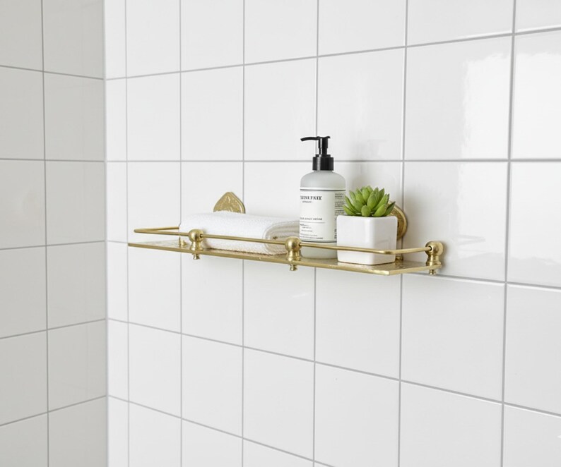 Puede incluir: Estante de ba&ntilde;o de metal dorado montado en azulejos blancos. El estante contiene una toalla blanca enrollada, una botella de jab&oacute;n y una peque&ntilde;a planta suculenta en maceta. El estante tiene detalles decorativos y una barra.