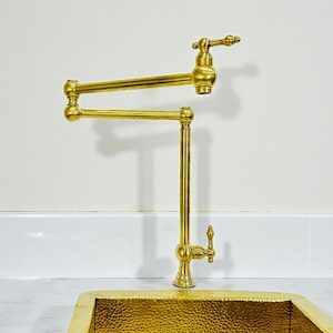 May include: A gold-coloured, wall-mounted pot filler tap with a swing arm design. The tap has a vertical pipe and a horizontal arm that extends and retracts. The tap is made of brass and has a classic design.