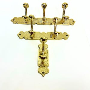 Pode incluir: Tr&ecirc;s cabides dourados com v&aacute;rios ganchos. Cada cabide tem um design decorativo e texturizado. Os ganchos s&atilde;o em forma de bola. Os cabides s&atilde;o montados em um fundo branco.