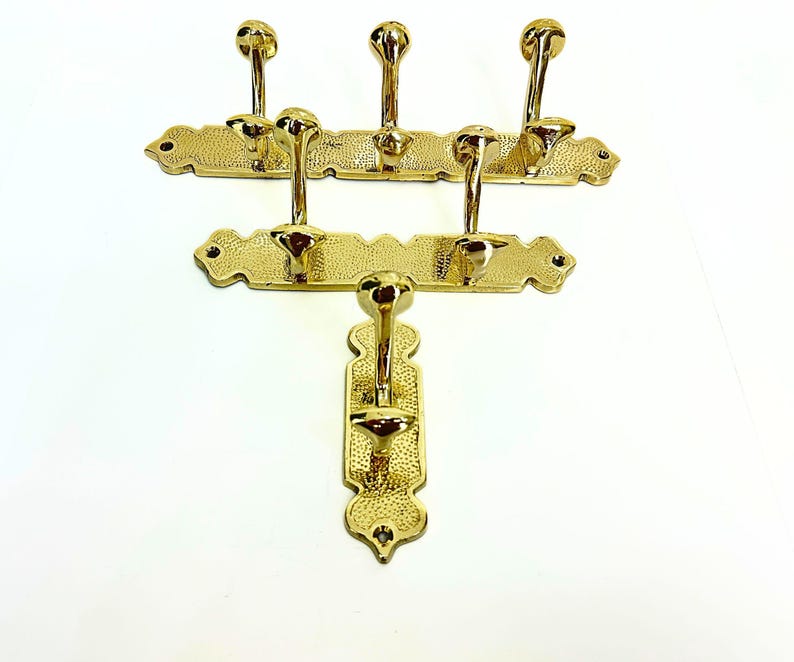 Pode incluir: Tr&ecirc;s cabides dourados com v&aacute;rios ganchos. Cada cabide tem um design decorativo e texturizado. Os ganchos s&atilde;o em forma de bola. Os cabides s&atilde;o montados em um fundo branco.