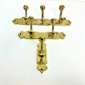 Pode incluir: Tr&ecirc;s cabides de lat&atilde;o com v&aacute;rios ganchos. Os dois suportes superiores t&ecirc;m cinco ganchos cada, e o suporte inferior tem dois. Os ganchos s&atilde;o arredondados e os suportes t&ecirc;m um design decorativo.