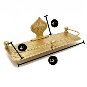 Puede incluir: Una estanter&iacute;a rectangular de color lat&oacute;n con un dise&ntilde;o grabado y ornamentado. La estanter&iacute;a mide 30 cm de largo y 10 cm de ancho, con una pieza trasera decorativa y detalles redondeados. La estanter&iacute;a mide 10 cm de alto. El dise&ntilde;o es probablemente para ba&ntilde;o o cocina.