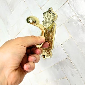 Pode incluir: Um gancho de parede de metal dourado com um design ornamentado. O gancho tem um bot&atilde;o arredondado e uma placa traseira decorativa com um padr&atilde;o texturizado. O gancho est&aacute; sendo segurado por uma m&atilde;o.