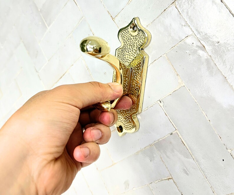 Pode incluir: Um gancho de parede de metal dourado com um design ornamentado. O gancho tem um bot&atilde;o arredondado e uma placa traseira decorativa com um padr&atilde;o texturizado. O gancho est&aacute; sendo segurado por uma m&atilde;o.