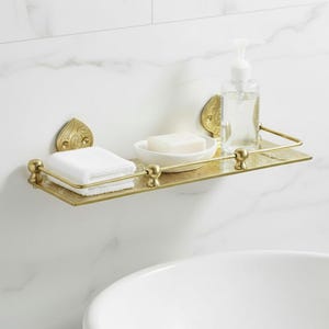 Puede incluir: Estante de ba&ntilde;o de metal dorado con detalles ornamentados. Contiene una pila de toallas blancas, una pastilla de jab&oacute;n en un plato blanco y un dispensador de jab&oacute;n l&iacute;quido transparente. Montado en una pared blanca con vetas de m&aacute;rmol.