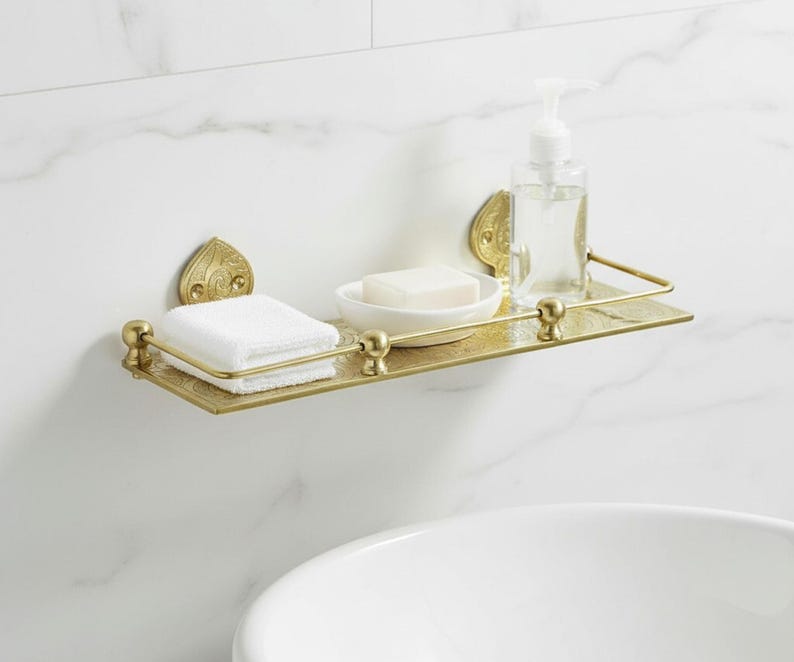 Puede incluir: Estante de ba&ntilde;o de metal dorado con detalles ornamentados. Contiene una pila de toallas blancas, una pastilla de jab&oacute;n en un plato blanco y un dispensador de jab&oacute;n l&iacute;quido transparente. Montado en una pared blanca con vetas de m&aacute;rmol.