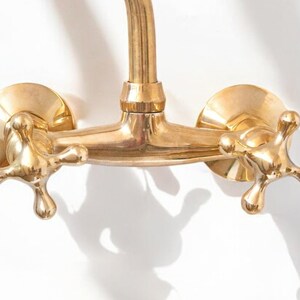 May include: A gold-toned brass faucet with two cross-shaped handles. The faucet is mounted on a wall and has a curved spout.
