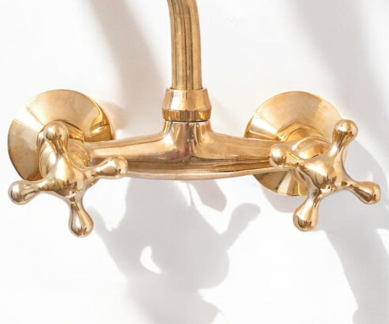 May include: A gold-toned brass faucet with two cross-shaped handles. The faucet is mounted on a wall and has a curved spout.