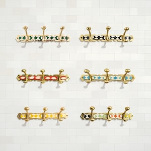 Op de afbeelding: Een verzameling decoratieve messing kapstokken met elk drie haken. De kapstokken hebben kleurrijke geometrische patronen in groen, zwart, rood, blauw, geel en oranje. Ze zijn bevestigd aan een witte bakstenen muur.