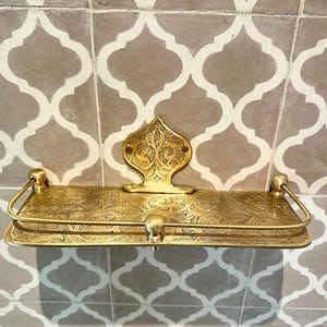 Puede incluir: Estante de lat&oacute;n ornamentado con un dise&ntilde;o floral en relieve. El estante tiene una placa trasera decorativa y un riel frontal curvo. El estante est&aacute; montado en una pared de azulejos estampados. El estante mide unos 30 cm de ancho.