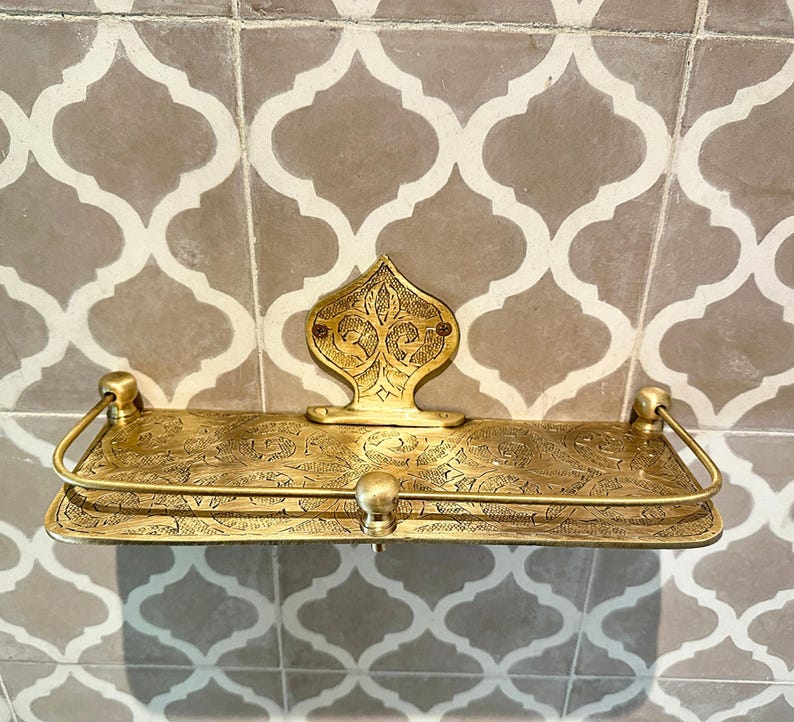 Puede incluir: Estante de lat&oacute;n ornamentado con un dise&ntilde;o floral en relieve. El estante tiene una placa trasera decorativa y un riel frontal curvo. El estante est&aacute; montado en una pared de azulejos estampados. El estante mide unos 30 cm de ancho.